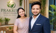 A man in a navy blue suit and a woman in a light pink sari stand smiling in front of a wall with the 'PRAKriti Healthcare' logo. The background features indoor plants, shelves, and wellness bottles.
