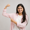 A smiling young woman with long dark hair poses with her arm bent, flexing her bicep while wearing a light pink button-up shirt over a white t-shirt, set against a solid light gray background.